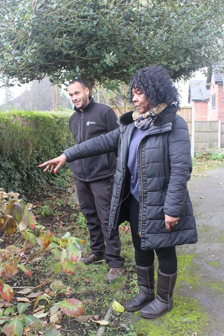 a man and a woman stand in a garden, the woman is pointing towards plants near the hedge